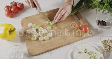 女人用<strong>一把</strong>刀在一张白色的厨房桌子上的木板上切下绿色韭菜做沙拉准备。 顶部视图
